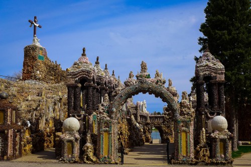 A decorative archway made of shells and stones, leading to a unique, artistic structure under a blue sky.