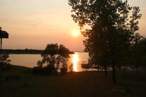 Sunset over a calm lake, with trees in silhouette and a boat docked along the shore.