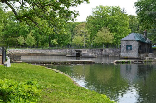 A serene park scene featuring a stone building by a calm pond, surrounded by lush green trees and grass.