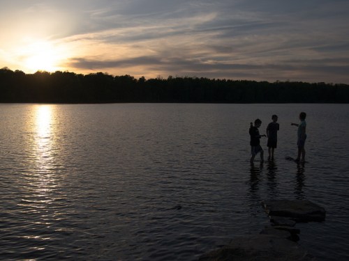 Three people stand in shallow water at sunset, with a serene lake and trees in the background.