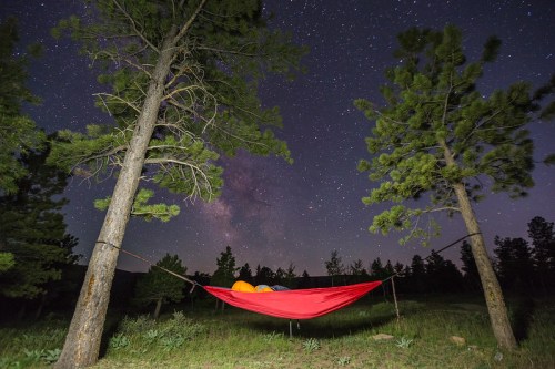 A red hammock suspended between two trees under a starry night sky, with the Milky Way visible in the background.