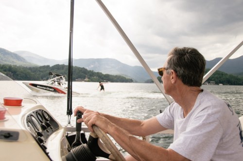 A man steers a boat on a lake, watching a person water skiing in the distance against a backdrop of mountains.
