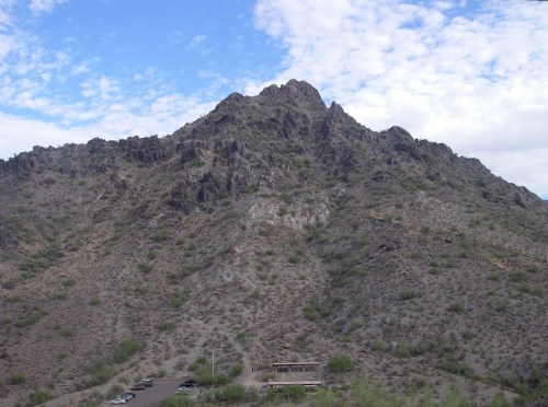 A rocky mountain peak rises above a desert landscape under a partly cloudy sky. Sparse vegetation is visible.