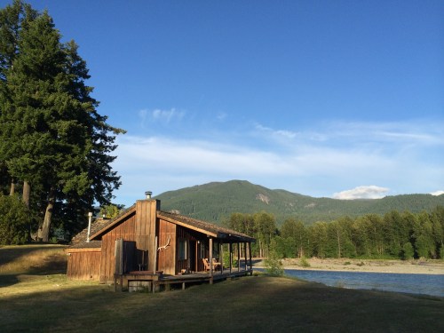 A rustic wooden cabin by a river, surrounded by trees and mountains under a clear blue sky.
