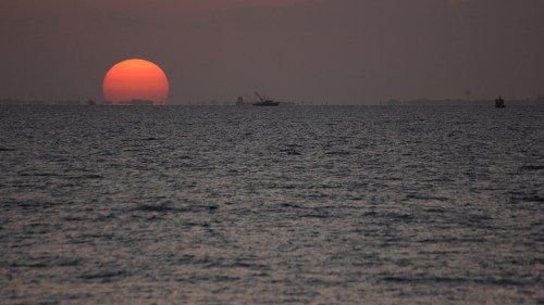 A large orange sun sets over a calm sea, with silhouettes of boats in the distance against a dusky sky.