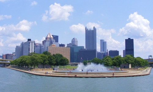 City skyline of Pittsburgh with a fountain, green park area, and blue sky with clouds.