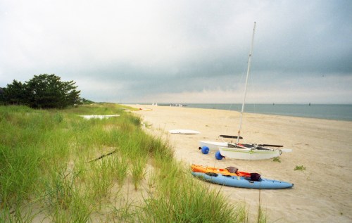 A sandy beach with grass, kayaks, and sailboats under a cloudy sky.