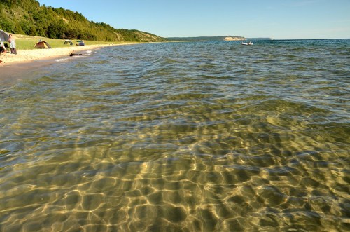 A serene beach scene with clear water, gentle waves, and green trees lining the shore under a blue sky.
