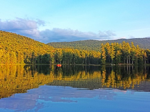 A serene lake surrounded by lush green mountains, reflecting the sky and trees, with a small red canoe in the distance.