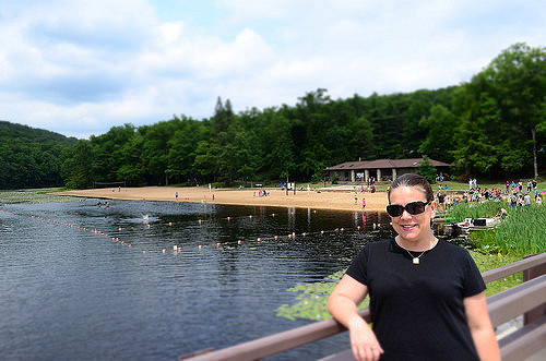 A woman in sunglasses stands by a lake with a sandy beach and people enjoying the outdoors in the background.