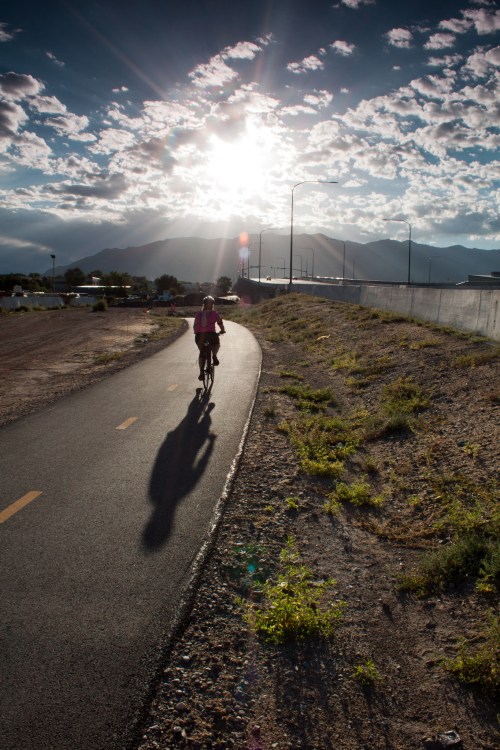A cyclist rides along a path with mountains in the background, illuminated by a dramatic sunset and scattered clouds.