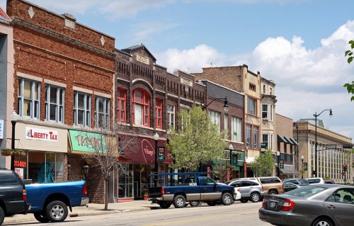 A street view featuring a row of shops and businesses with parked cars and trees under a partly cloudy sky.