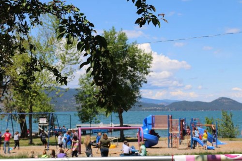 A playground by a lake, with swings and slides, surrounded by trees and people enjoying the sunny day.