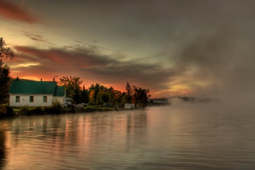 A serene lakeside scene at sunset, featuring a white house, mist over the water, and colorful clouds in the sky.