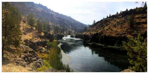 A serene river flows through a rocky landscape, surrounded by trees and hills under a clear sky.