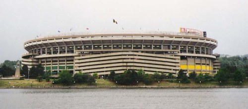 A large stadium with multiple levels, surrounded by trees and a body of water, under a cloudy sky.