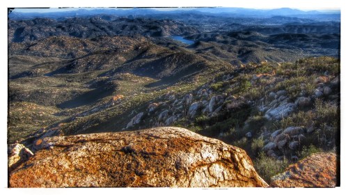 A panoramic view of rugged mountains and valleys, with rocky terrain and a distant lake under a clear sky.