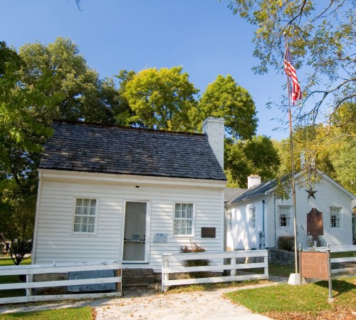 Historic white house with a black roof, surrounded by trees, featuring an American flag and a nearby informational sign.