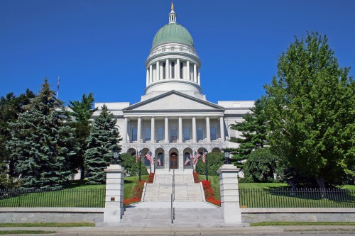 The image shows a grand building with a green dome, flanked by trees and flags, under a clear blue sky.