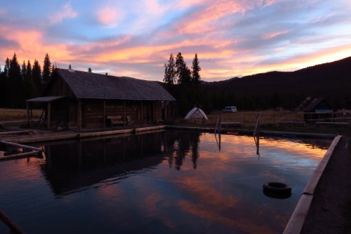 A serene landscape at sunset, featuring a rustic cabin by a reflective pond surrounded by trees and mountains.