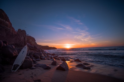 A serene beach at sunset, with gentle waves, rocky shore, and a surfboard resting on the sand.