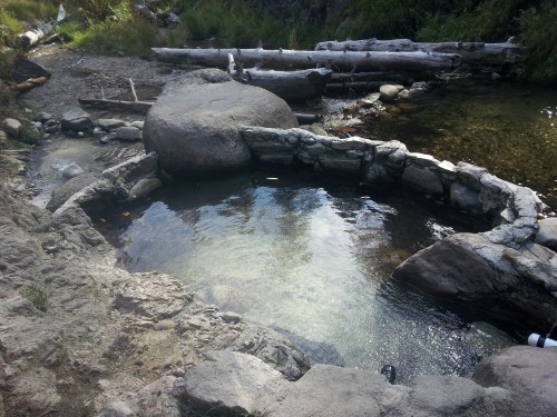 A natural hot spring surrounded by stone walls, with clear water and rocky terrain in a serene outdoor setting.