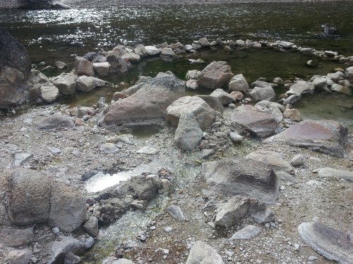 A rocky riverbank with scattered stones and a shallow, clear stream flowing through.