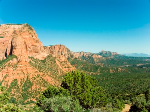 A panoramic view of red rock formations and green valleys under a clear blue sky.
