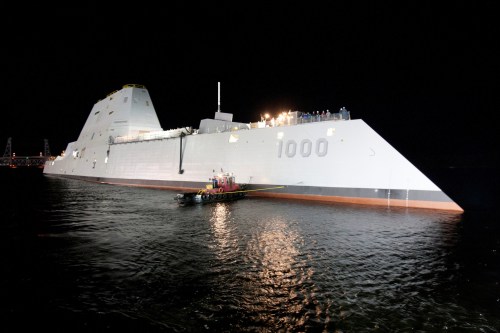 A large, modern naval ship illuminated at night, with a small tugboat alongside in calm waters.