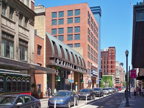 A busy city street featuring a theater, modern buildings, and parked cars under a clear blue sky.