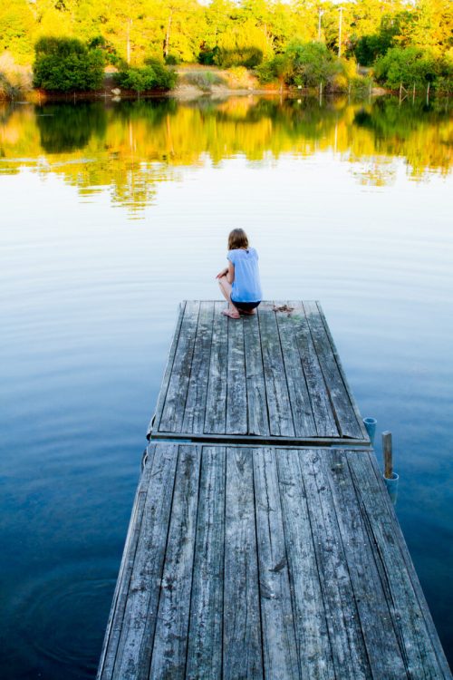 A child sits on a wooden dock, gazing at the calm water and surrounding greenery.