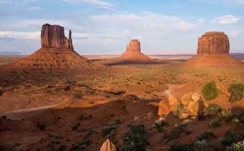 A scenic view of Monument Valley featuring three iconic sandstone buttes under a blue sky with scattered clouds.