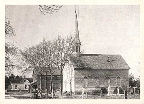 Black and white image of a wooden church with a tall steeple, surrounded by bare trees and a few nearby buildings.