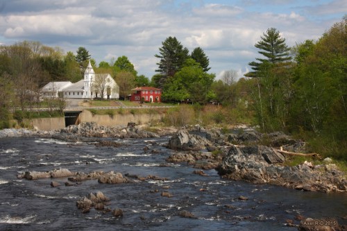 A scenic river flows past rocky banks, with a white church and red building nestled among green trees under a cloudy sky.