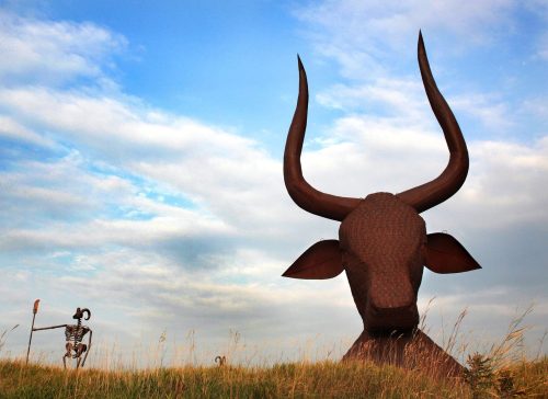 A large metal bull sculpture towers over a grassy field, with a skeleton figure holding a spear nearby under a cloudy sky.
