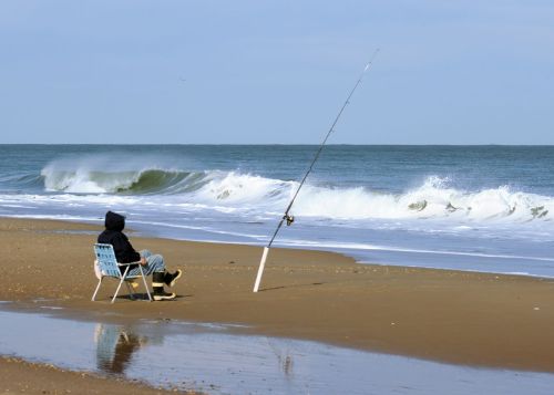 A person sits on a beach chair, fishing rod planted in the sand, with waves crashing in the background.