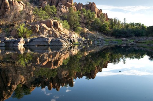 A serene landscape featuring rocky cliffs reflected in calm water, surrounded by trees and a clear blue sky.