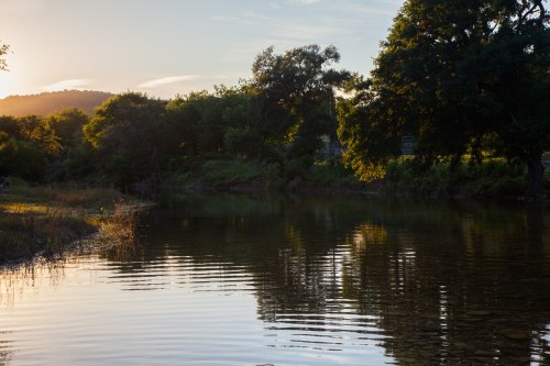 A serene river at sunset, with trees reflecting on the water's surface and gentle ripples.
