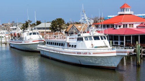 Two fishing boats docked by a waterfront building with a red roof, surrounded by calm water and clear blue skies.