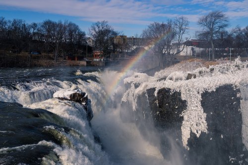 A waterfall cascades over icy rocks, with a rainbow arcing above and trees in the background under a clear blue sky.