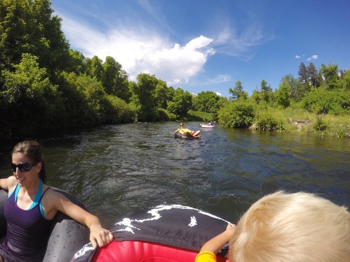 A sunny river scene with people floating on tubes, surrounded by lush greenery and blue skies.