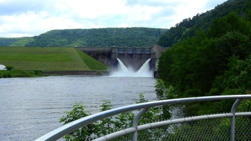 A dam releasing water into a river, surrounded by green hills and trees under a cloudy sky.