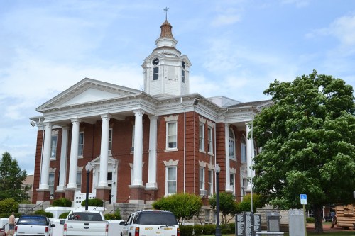 Historic brick courthouse with white columns and a clock tower, surrounded by trees and parked cars under a blue sky.