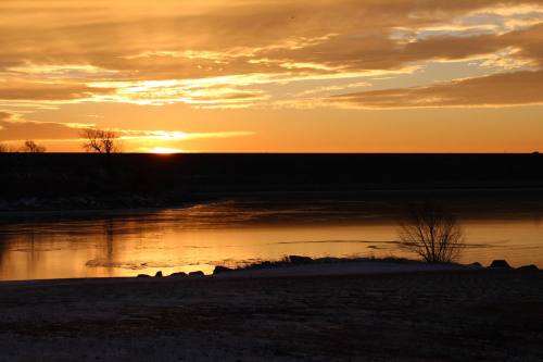 Sunset over a calm river, with golden hues reflecting on the water and silhouettes of trees along the shore.