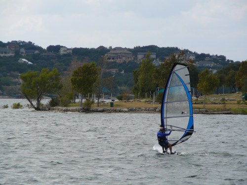 A person windsurfing on a lake, with trees and houses in the background under a cloudy sky.