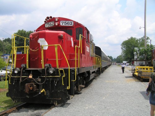 A red train engine parked on a gravel track, with a person walking nearby and trees in the background.