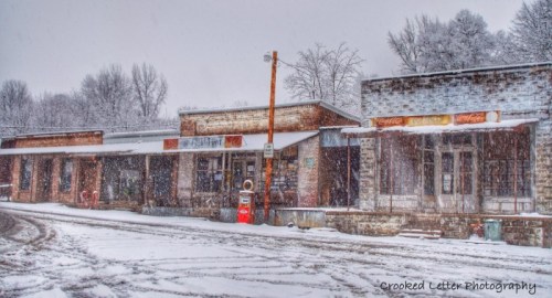 Snow-covered old storefronts line a quiet street, with falling snow creating a serene winter scene.