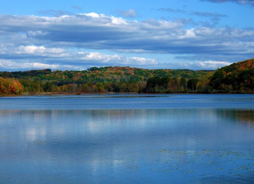 A serene lake reflects blue skies and colorful autumn foliage on the surrounding hills.