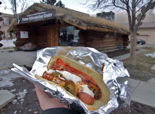 A hand holds a hot dog with toppings in front of a log cabin visitor information center.