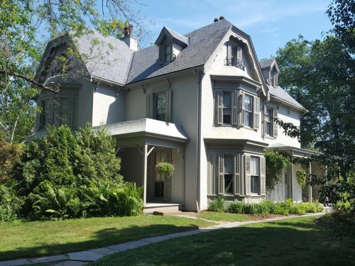 A charming two-story house with a gray roof, white walls, and lush greenery surrounding it on a sunny day.